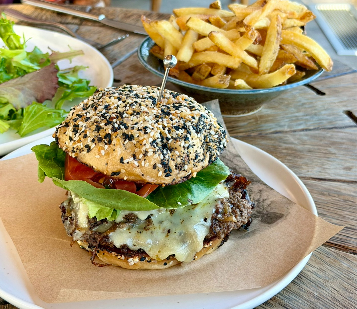 A sesame-seed burger with lettuce and tomato on a white plate, accompanied by a side of fries and a green salad on a wooden table.