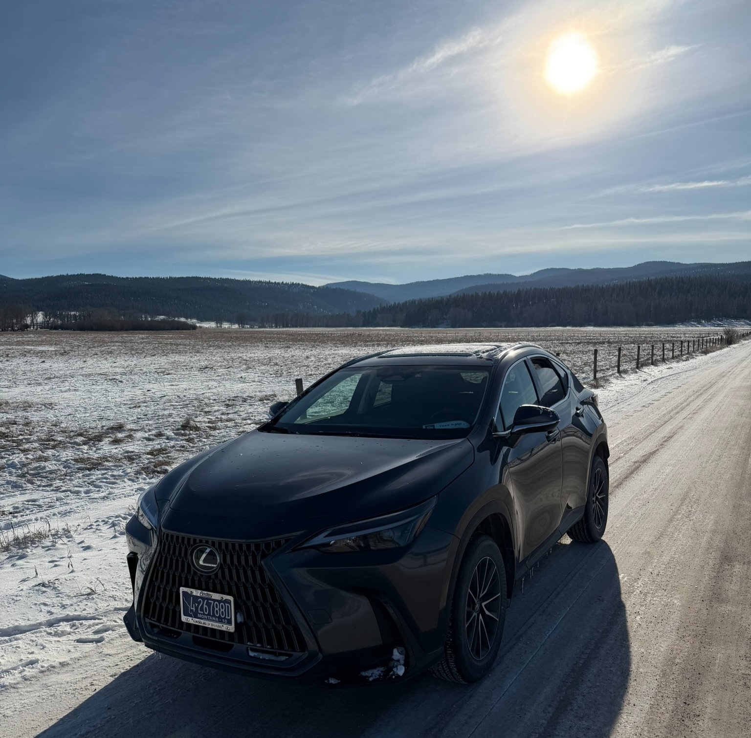 Black car on a snowy road under a bright sun, with mountains and a field in the background. A serene winter landscape.