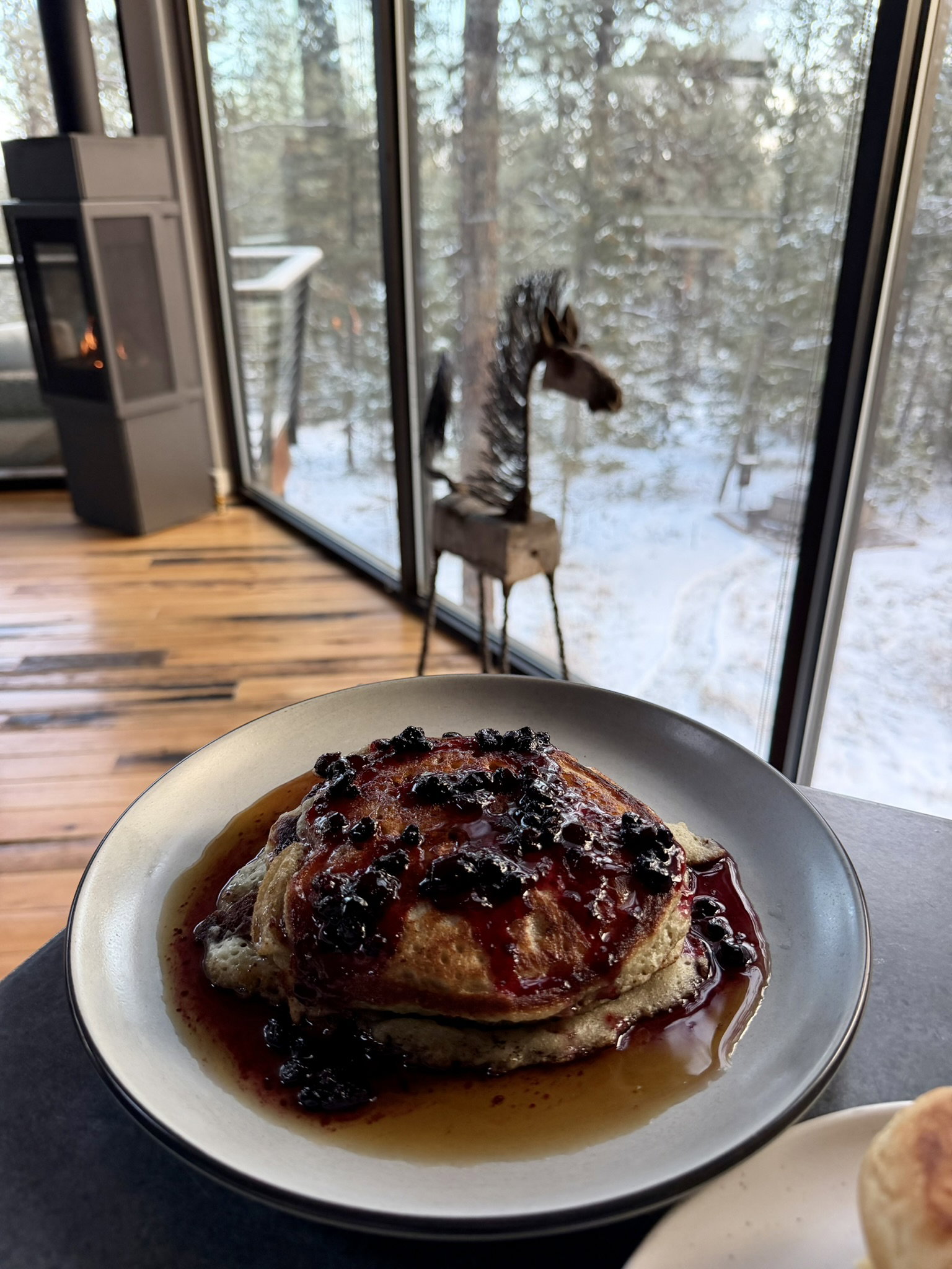Pancakes with syrup and berries on a table in a cozy cabin with a fireplace. Snowy forest visible through large windows, creating a warm vibe.