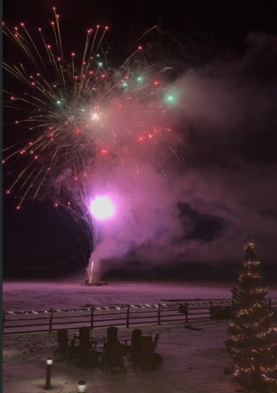 Fireworks burst in colorful sparks over a snowy landscape with a decorated Christmas tree and lit chairs nearby, creating a festive atmosphere.