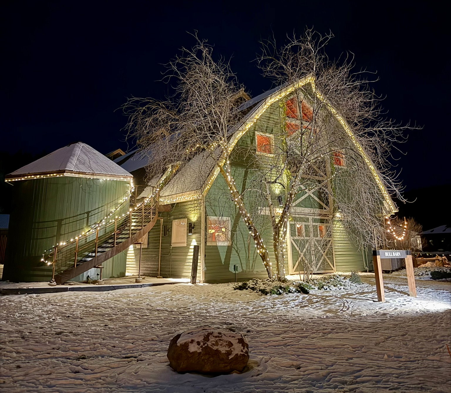 A snow-covered green barn with twinkling lights, bare trees, and a sign reading "Bull Barn" at night, creating a festive winter scene.
