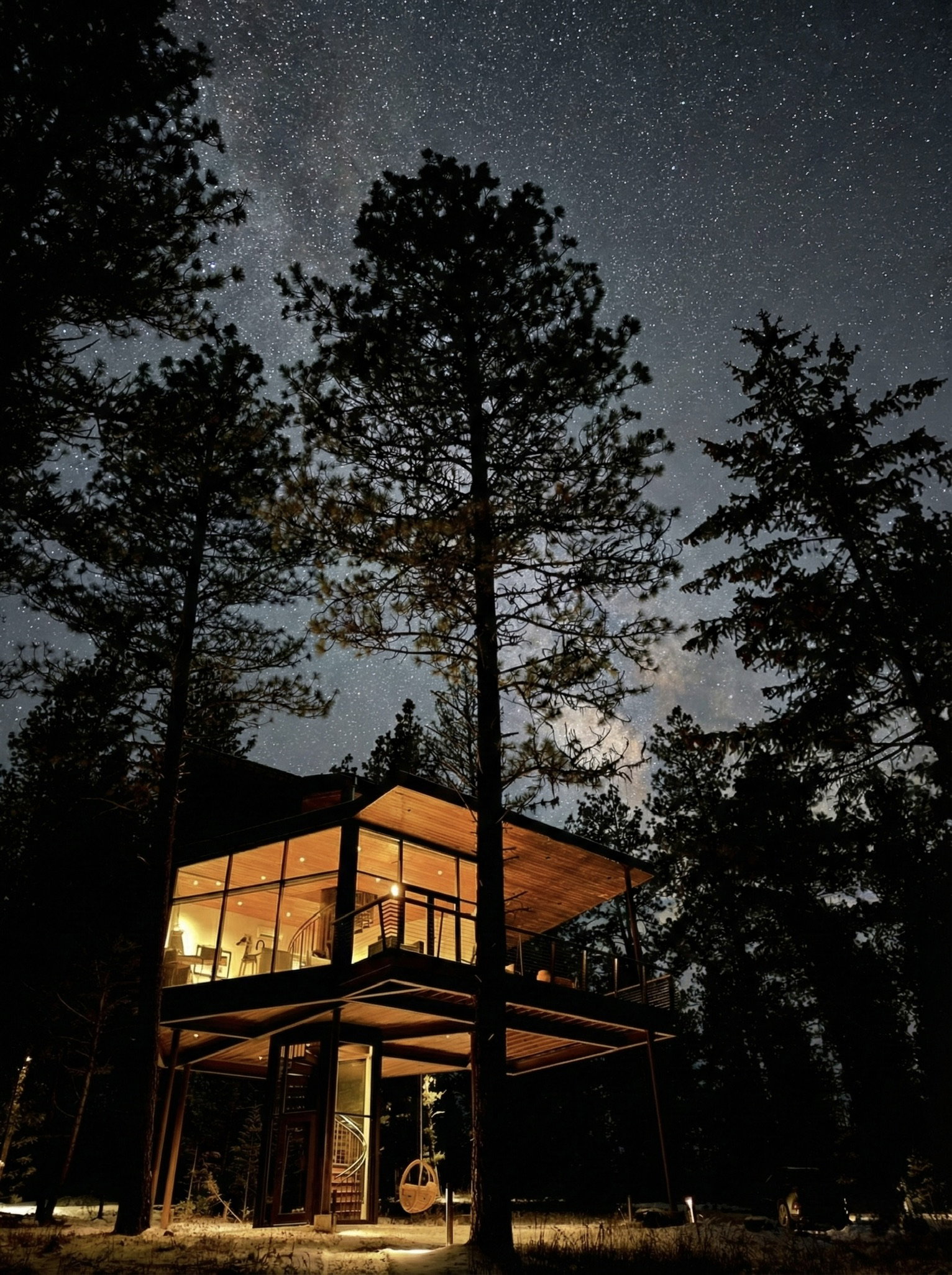 A modern, illuminated treehouse stands among dark trees under a starry night sky, creating a warm and serene atmosphere.