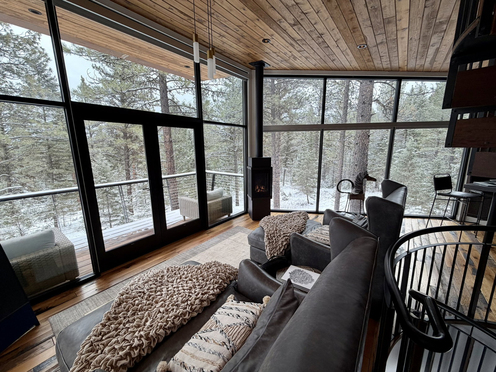 Cozy interior with dark sofas, patterned blankets, and a view of snowy pine trees through large windows. Wooden ceiling, spiral staircase.