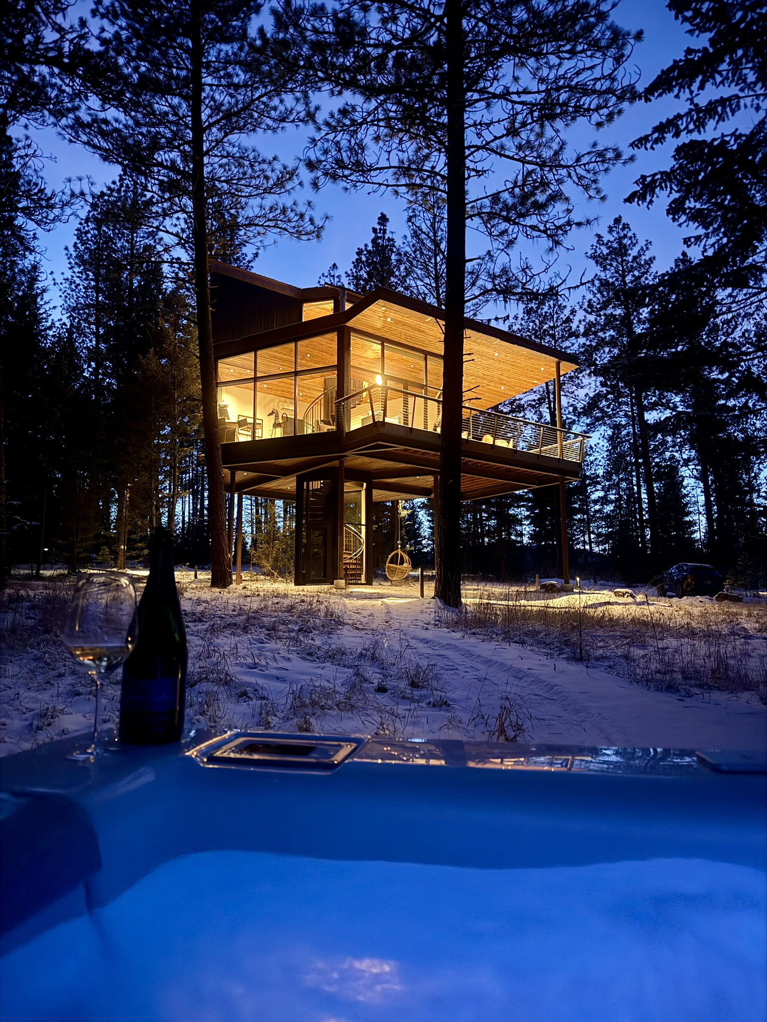 Modern cabin with warm lights in snowy forest at dusk, viewed from a hot tub with a wine bottle and glass. Peaceful and serene mood.