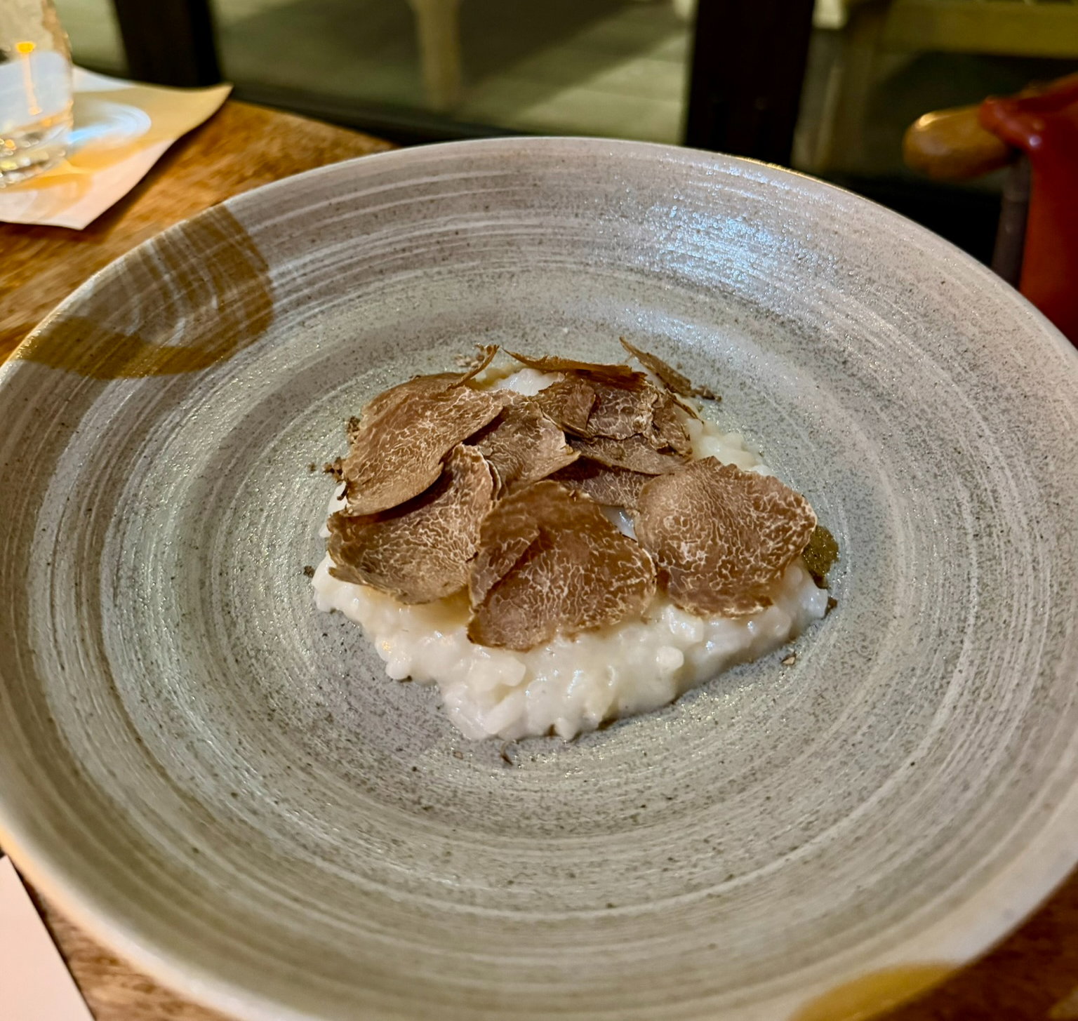 Truffle slices on creamy risotto served in a textured gray bowl, placed on a wooden table. Dimly lit, cozy dining ambiance.