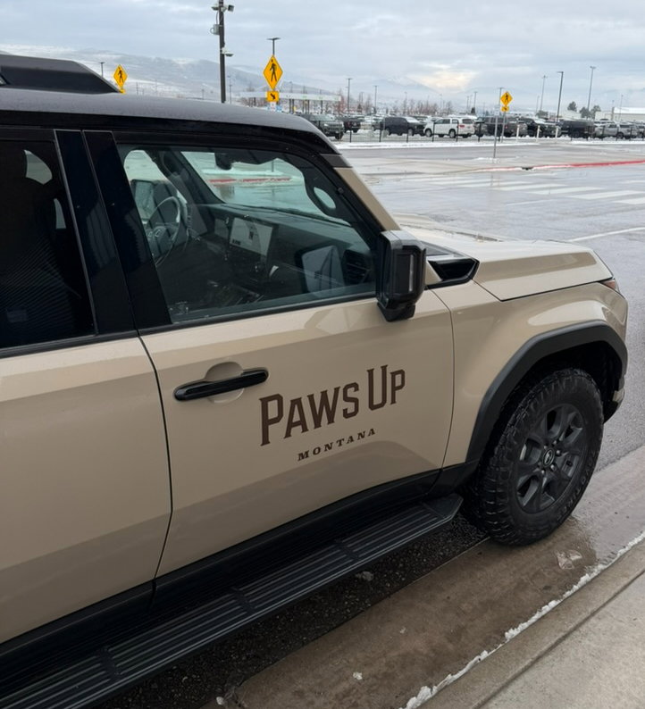 Beige SUV with "Paws Up Montana" on the side, parked near a snowy sidewalk. Overcast sky and a parking lot background.