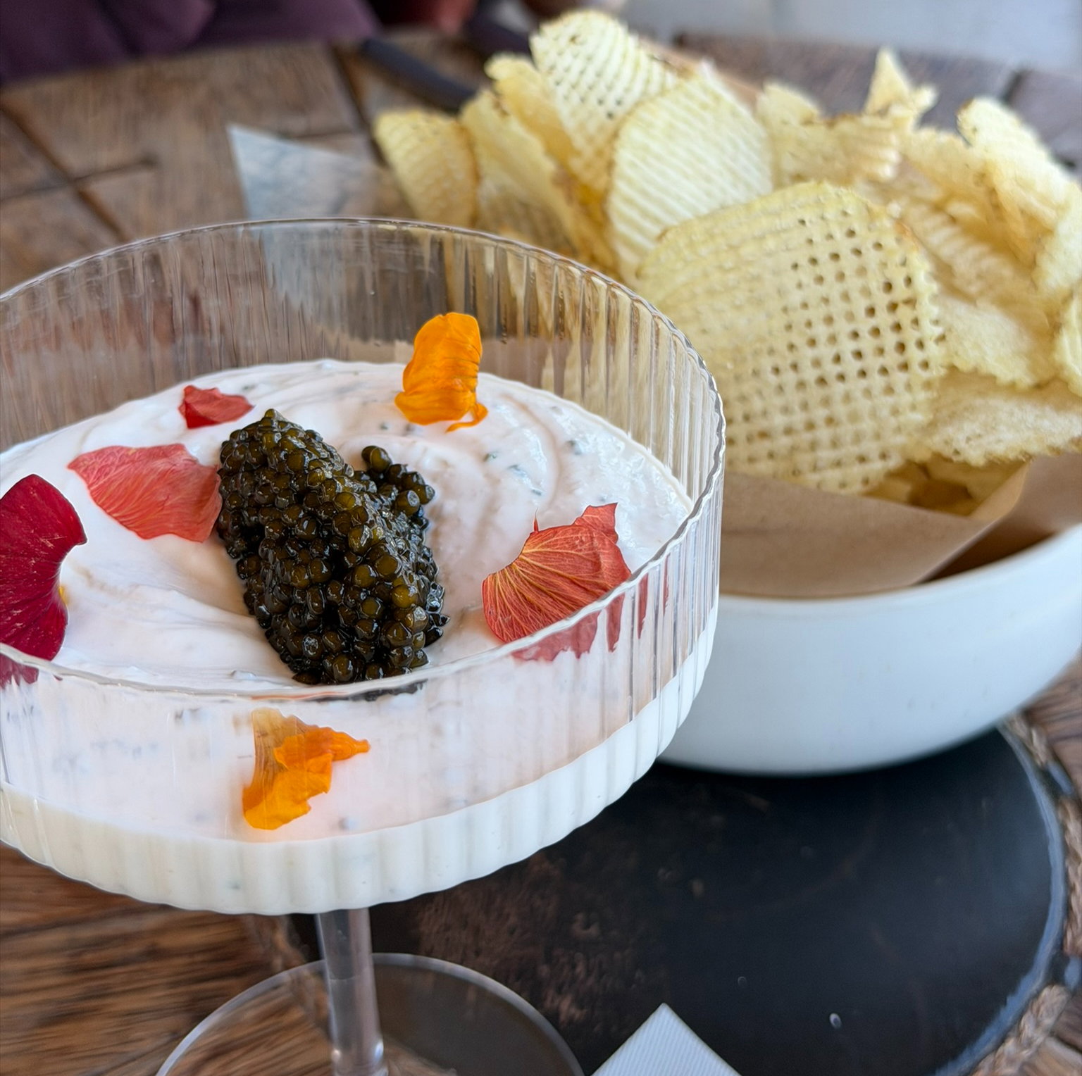 Glass bowl with creamy dip, topped with caviar and colorful petals, next to a bowl of waffle-cut potato chips on a wooden table.