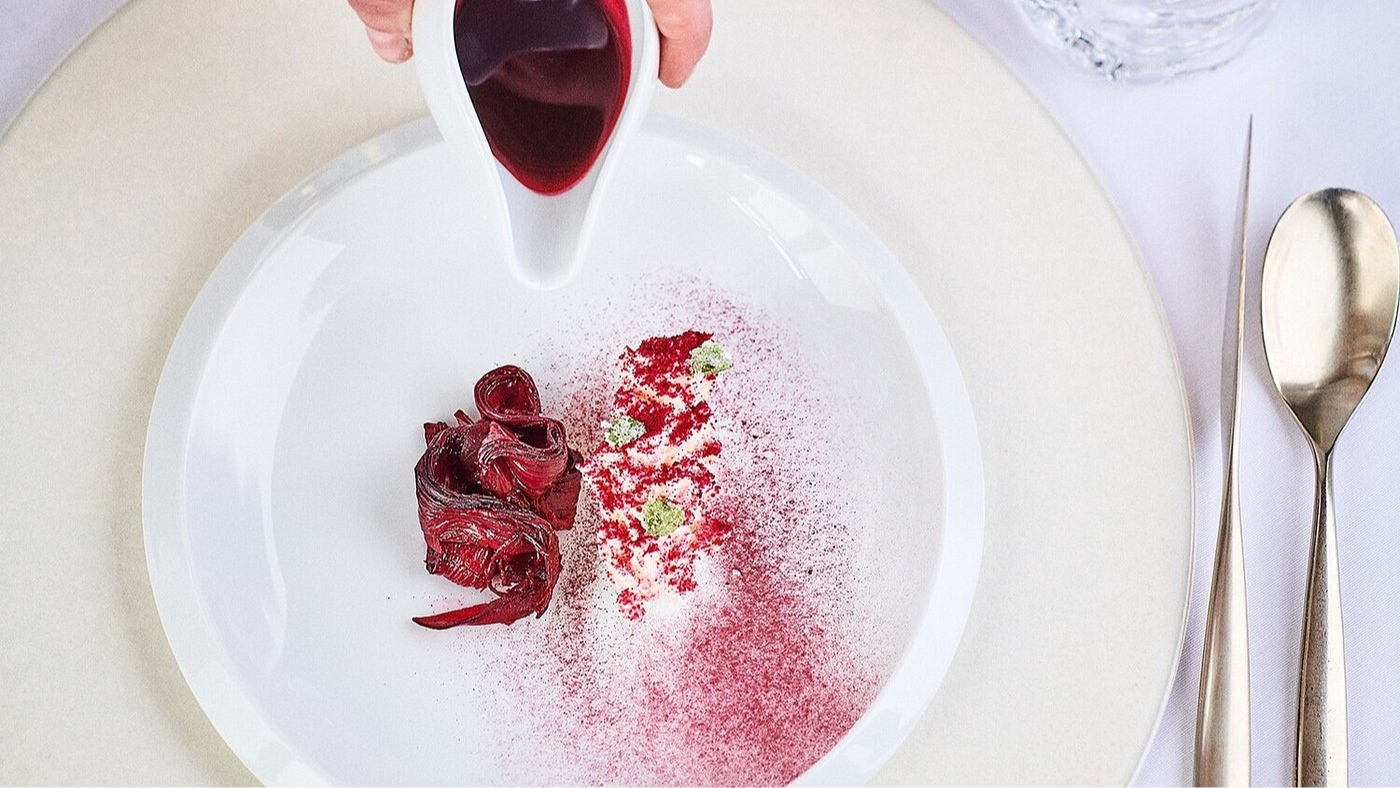 A hand pours a deep red sauce onto a white plate with red and green garnish. Silverware lies on a white tablecloth beside the plate.