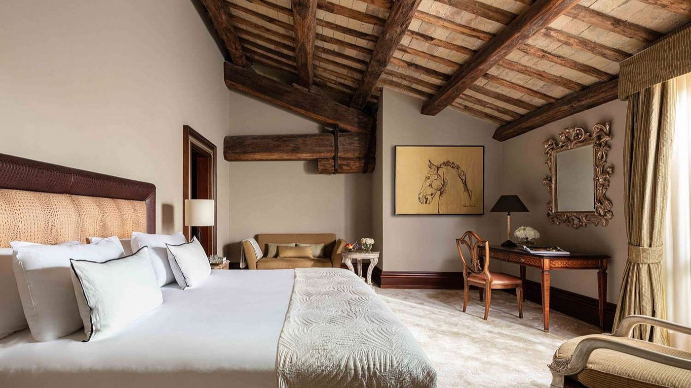 Cozy bedroom with a large bed, rustic wood beams, beige walls, and horse artwork. An elegant desk and chair beside a window with drapes.