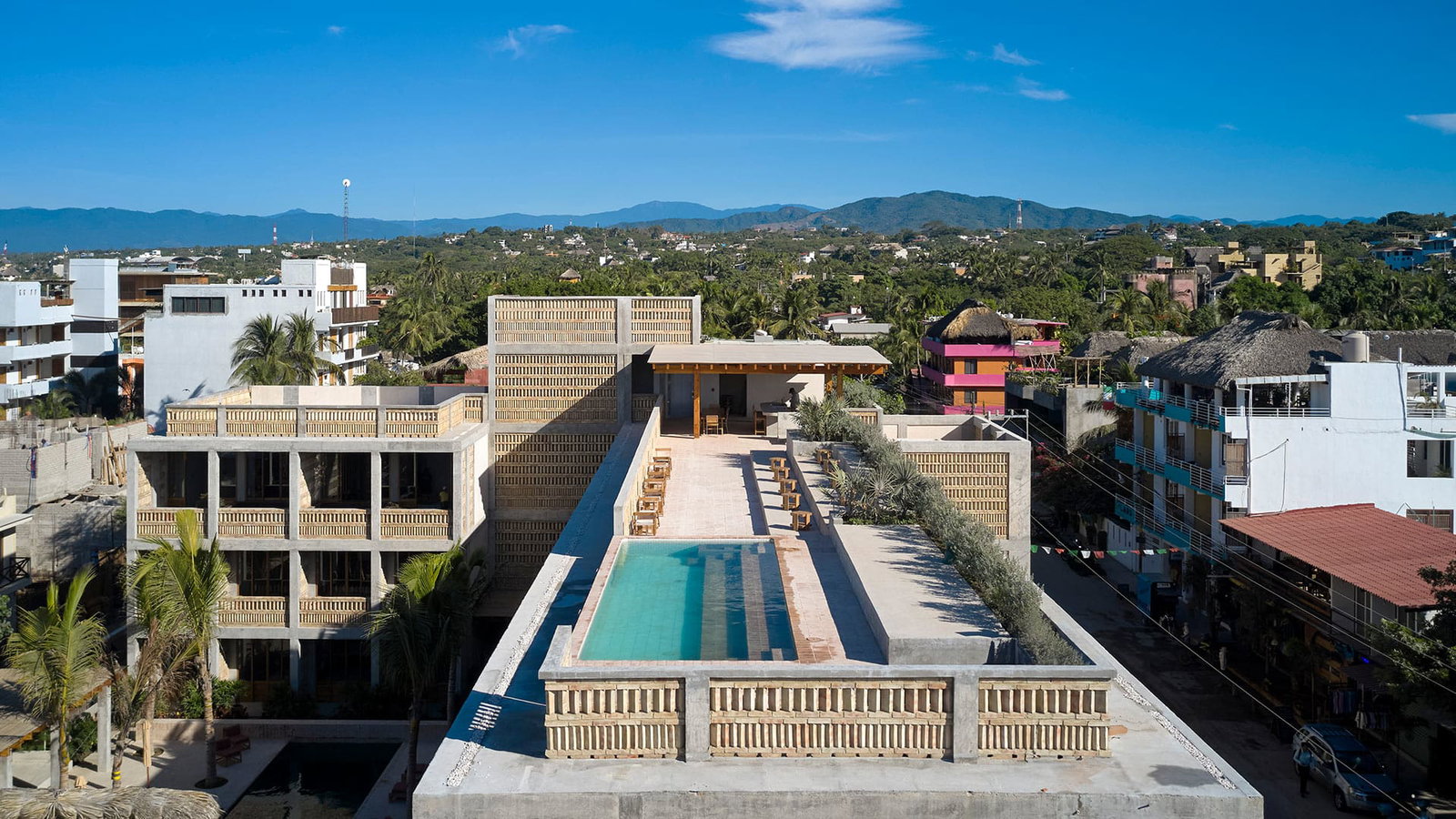 Aerial view of the rooftop pool and bar at Hotel Humano surrounded by palm trees. Colorful buildings and mountains are in the distant background.