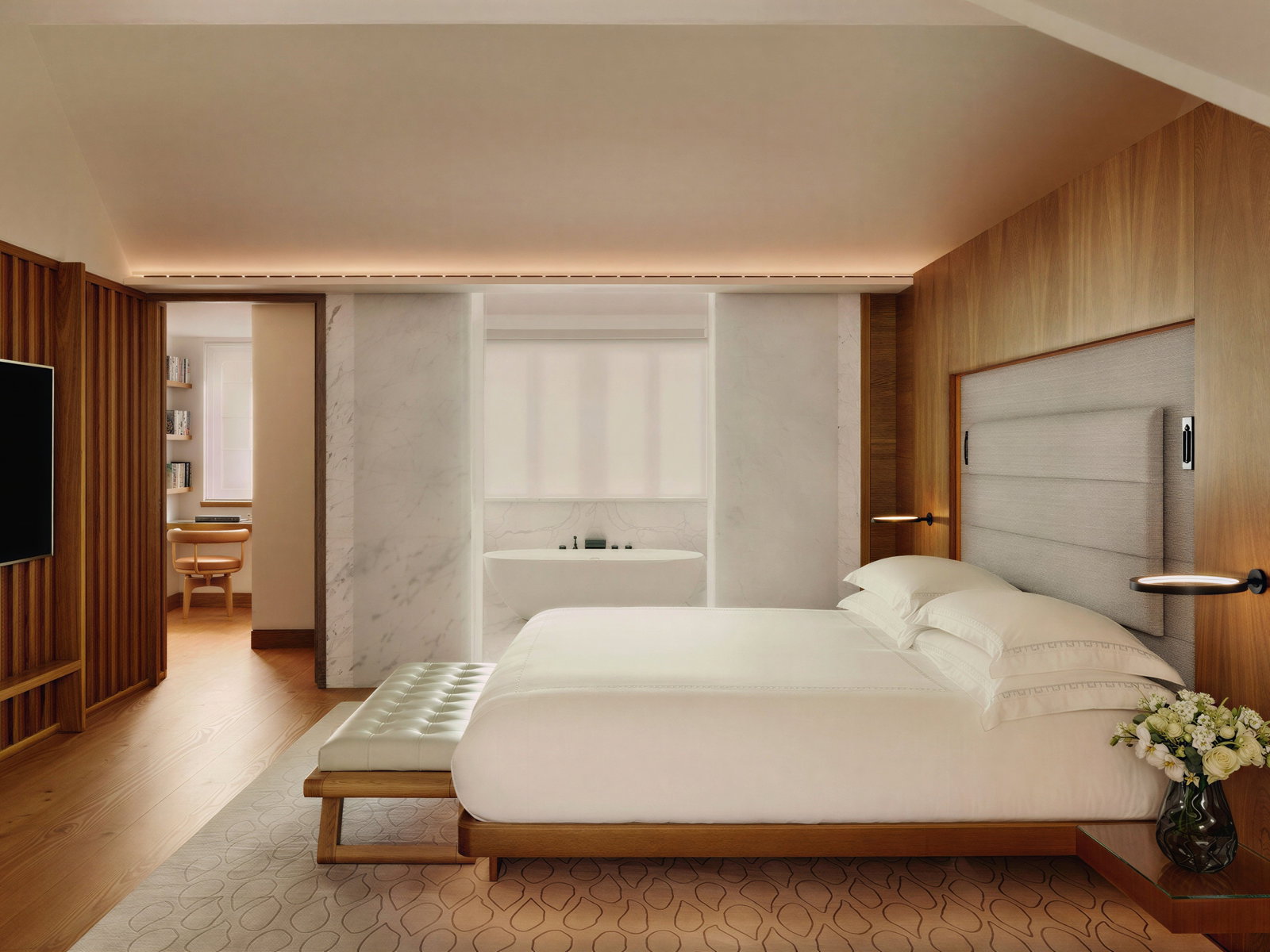Modern bedroom with a white bed, wood accents, and marble walls. A vase of flowers sits on the bedside table. Calm, minimalist vibe.