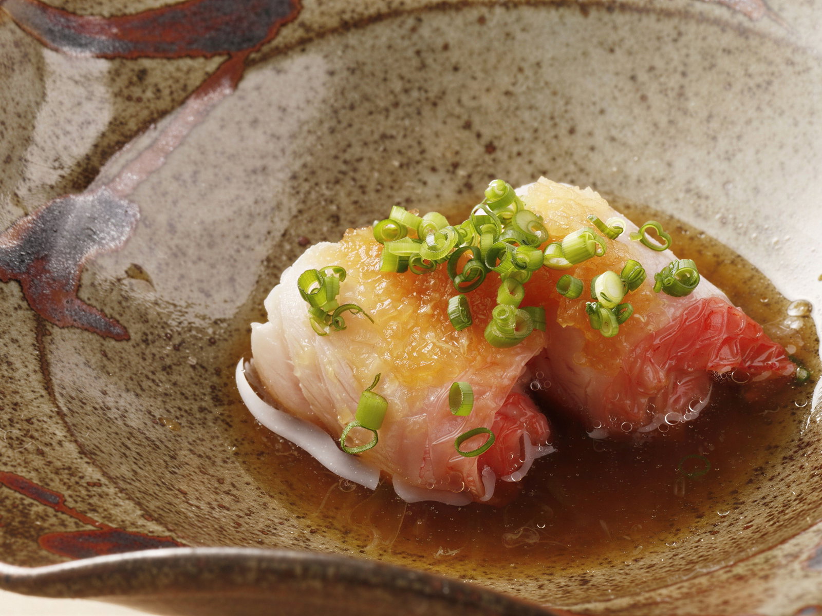 Sushi topped with green onions on a ceramic plate. Fish has a pink hue, surrounded by brown sauce, creating an appetizing look.