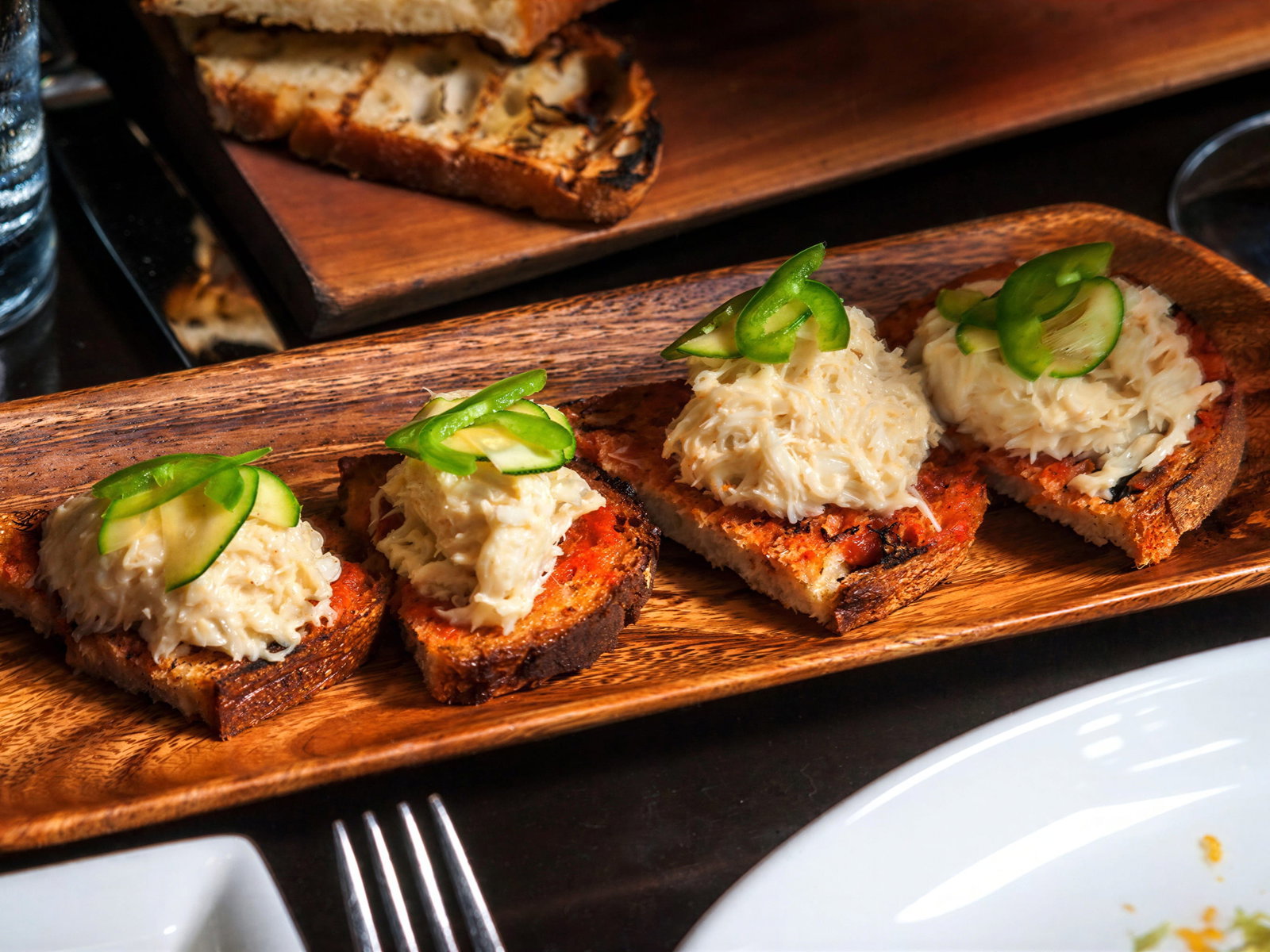 Blue crab crostini from Locanda verde with sliced jalapeno peppers on a wooden tray, in a cozy dining setting.