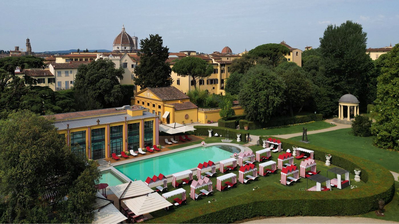 Rooftop view of a luxurious pool surrounded by red-and-white striped cabanas. Historic buildings and greenery are in the background.