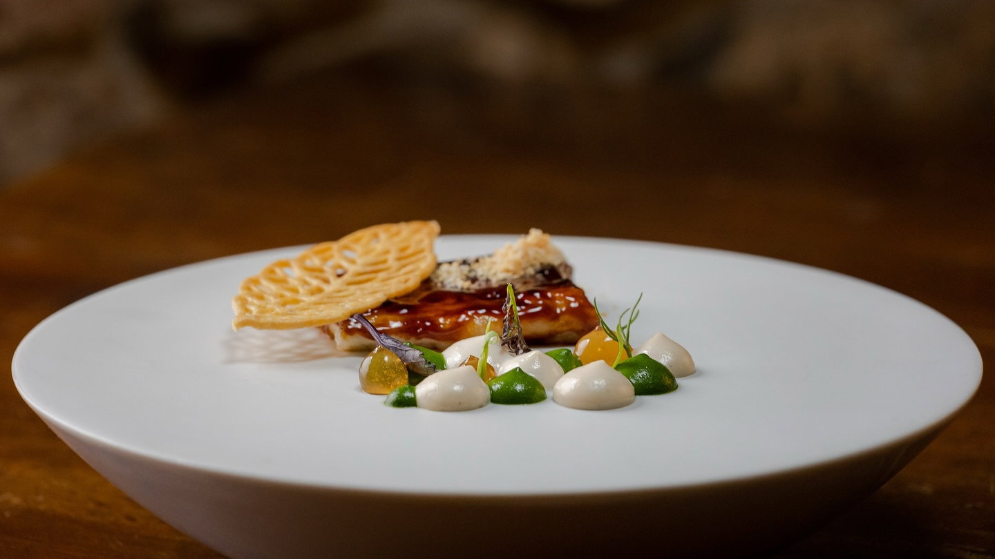Gourmet dish on a white plate with vibrant sauces, garnished with herbs and a lattice wafer. Set against a warm, blurred background.