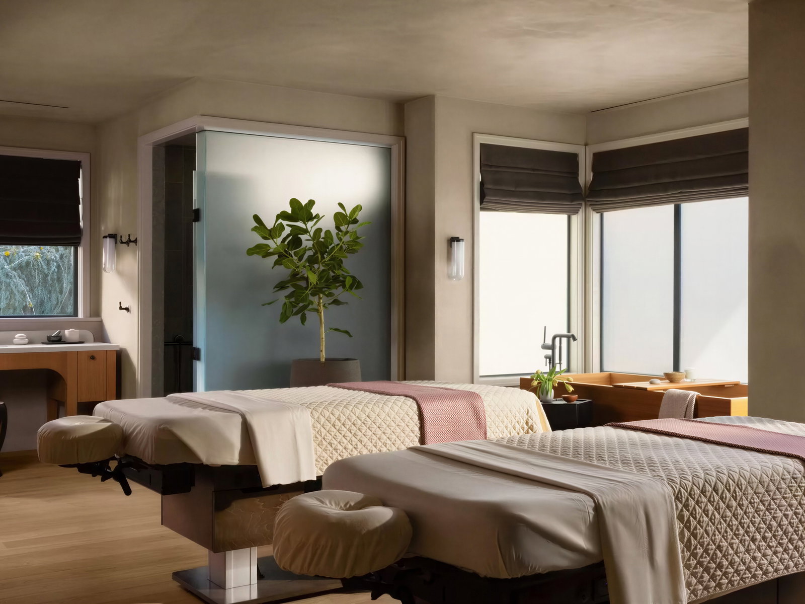 Calm spa room with two massage tables draped in white and pink blankets. A potted plant near frosted glass, wooden accents, serene ambiance.