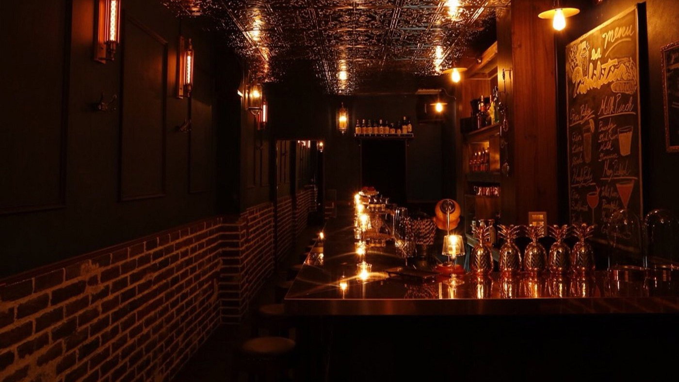 Dimly lit bar with ornate tin ceiling, glowing pendant lights, empty stools, and a chalkboard menu. Cozy and inviting atmosphere.