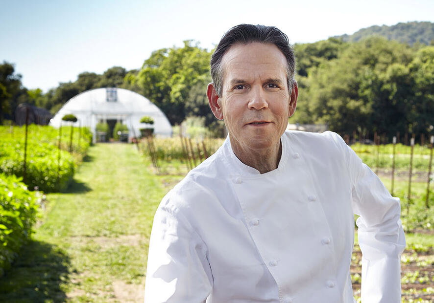 A man in a classic white chef's outfit poses amongst rows of fresh fruit and vegetables.