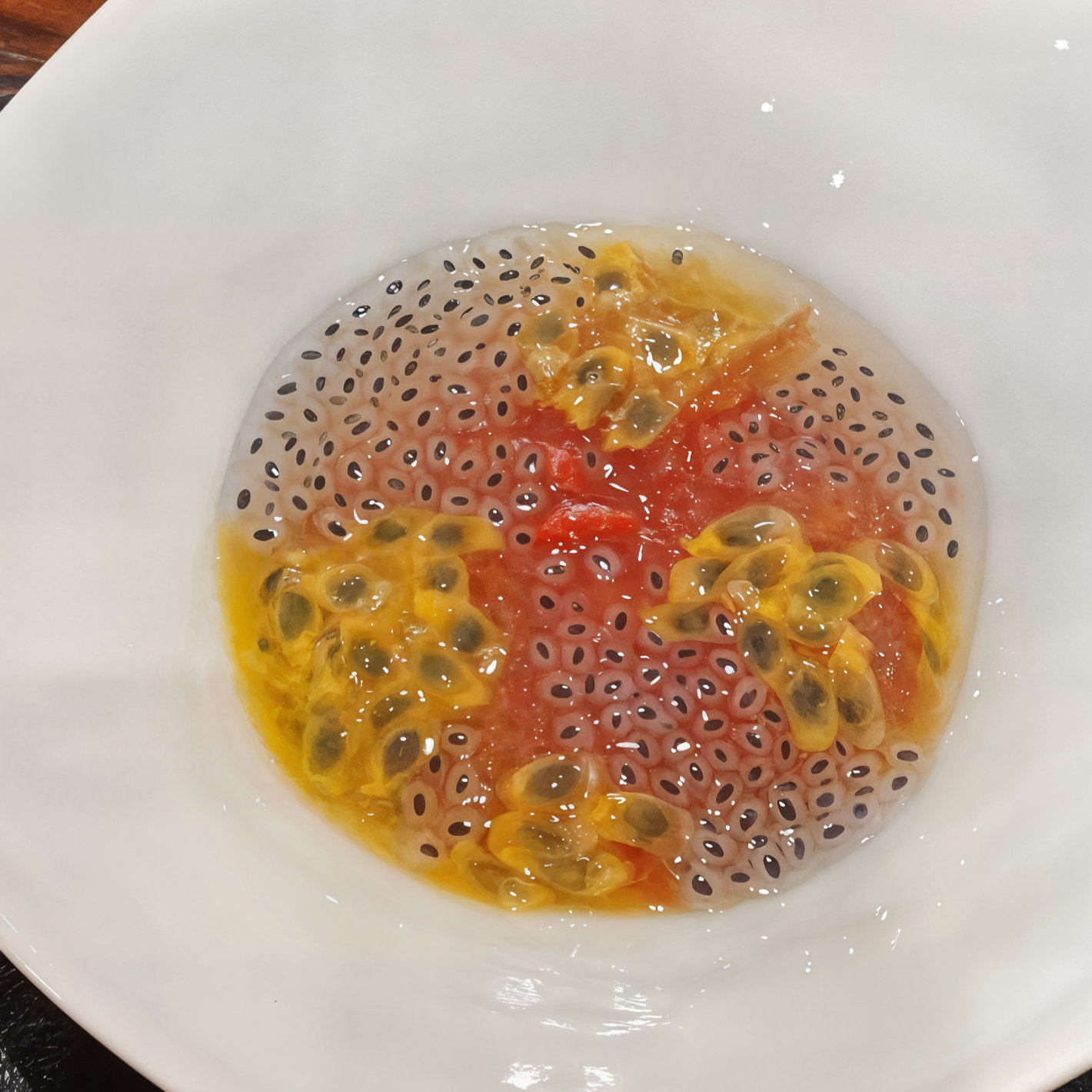 A white bowl with passion fruit seeds in gel, featuring black and red seeds in a clear, jelly-like liquid. Bright and colorful.