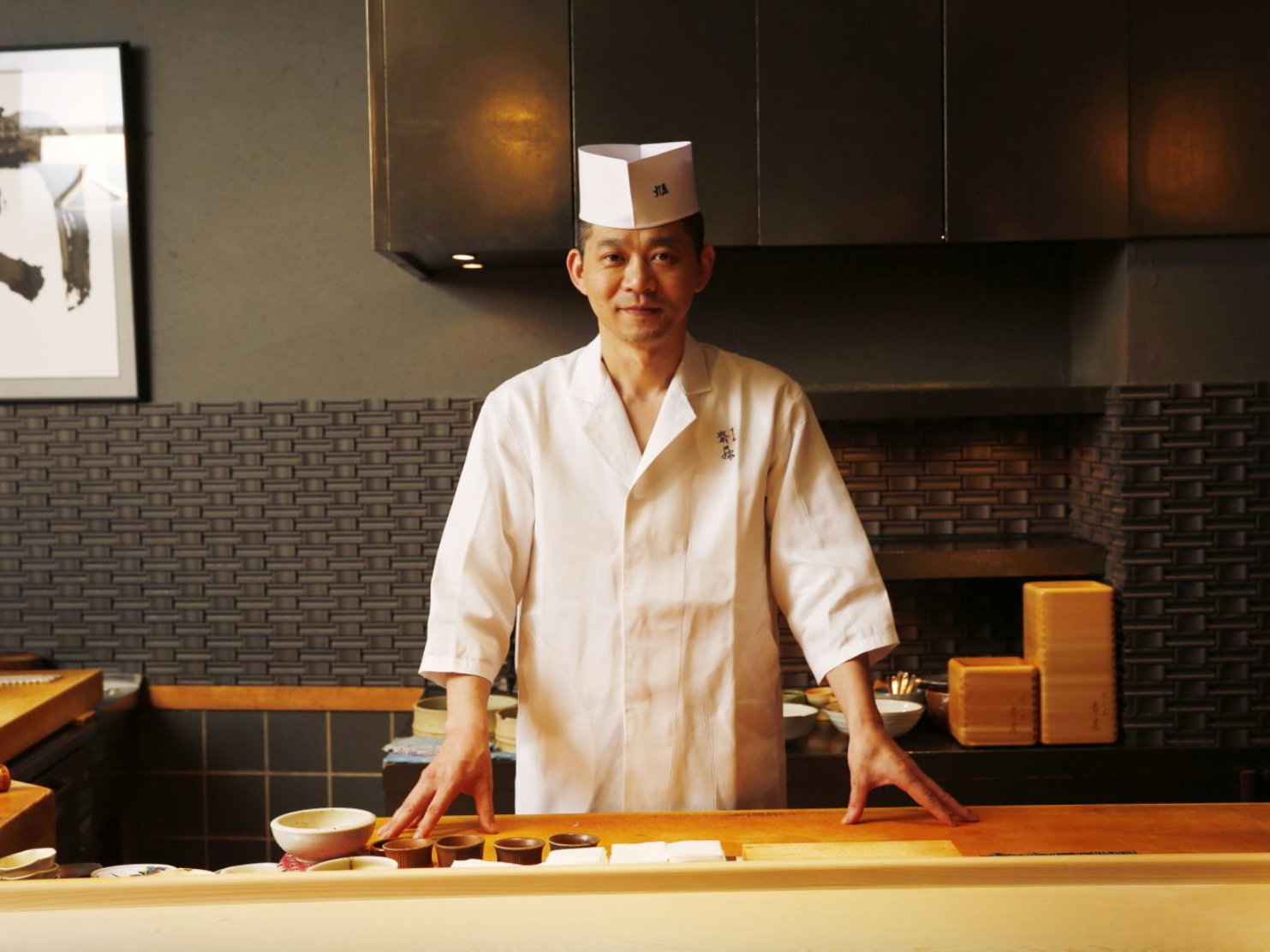 A sushi chef in a white uniform stands behind a wooden counter in a dimly lit restaurant. Bowls are on the counter, and a calm mood prevails.