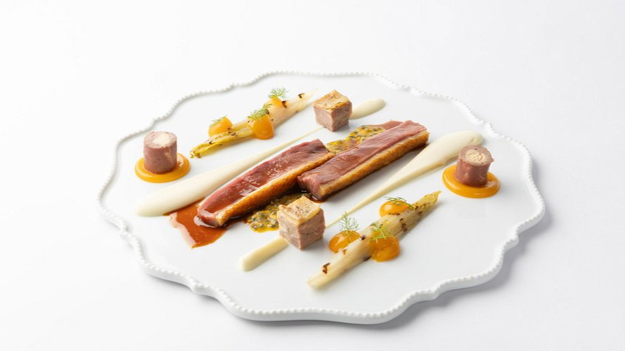 Gourmet dish on white plate: duck breast, cubes of meat, pureed swirls, and orange blobs, elegantly arranged with asparagus spears.