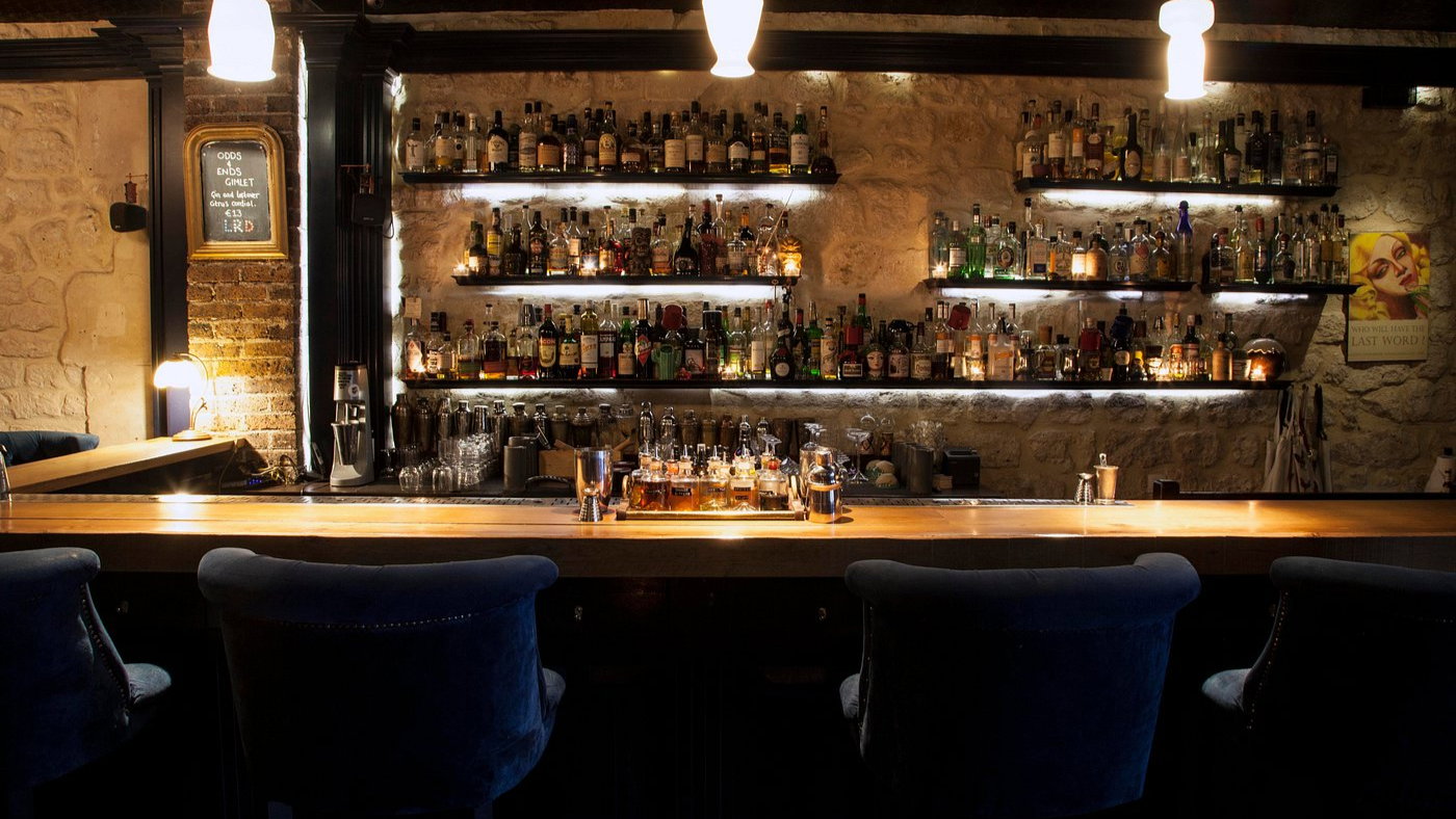 Cozy bar with velvet chairs, stone walls, and lit shelves full of liquor bottles. A sign reads "GIMLET." Warm, ambient lighting.