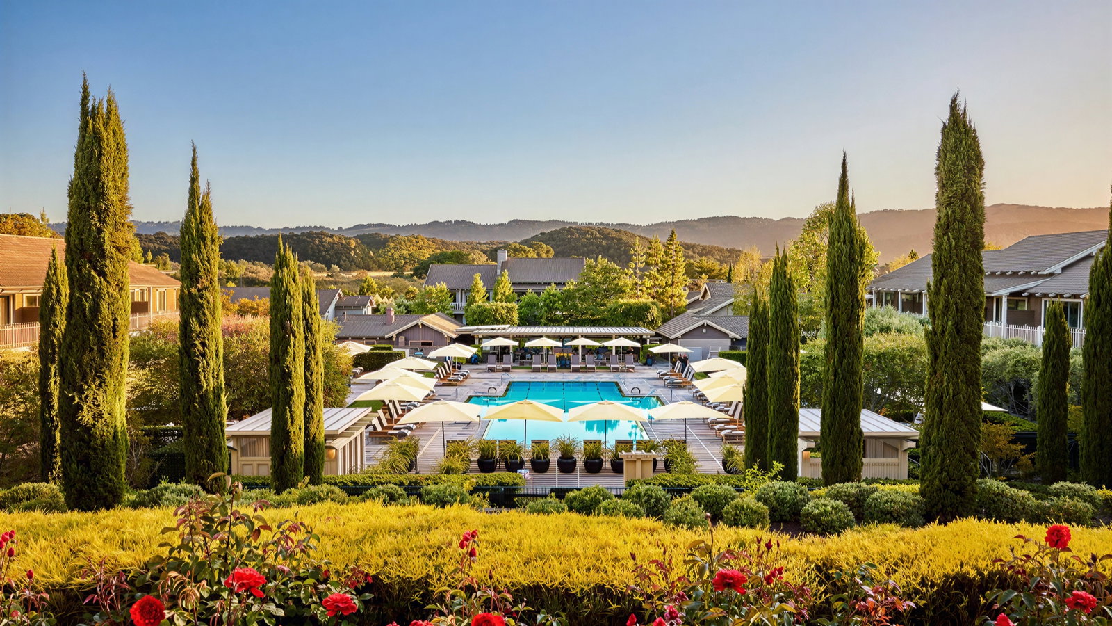 Luxurious pool area with sun umbrellas, surrounded by lush greenery and tall cypress trees, under a clear blue sky. Mountains in the background.