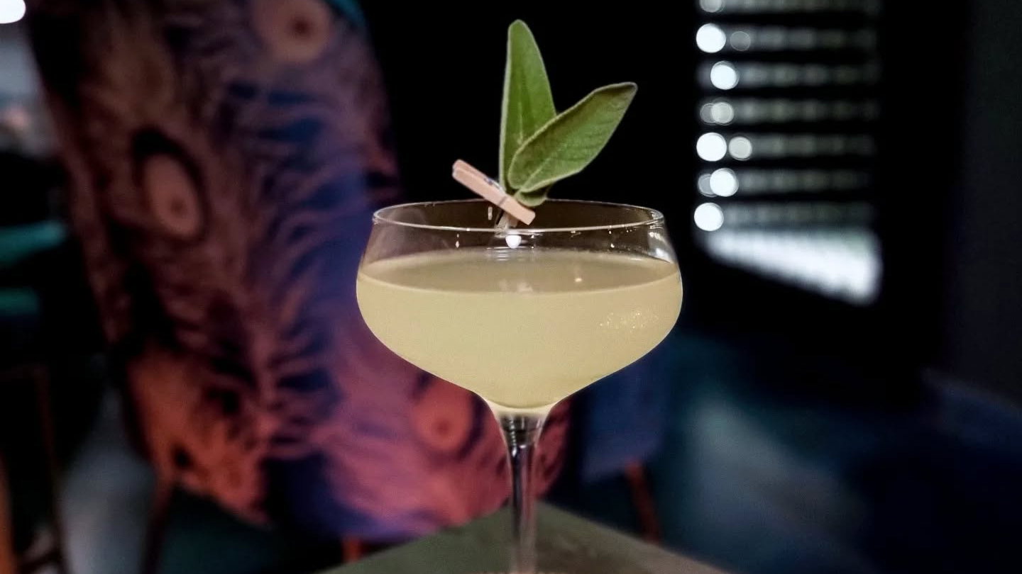 A yellow cocktail garnished with green leaves in a glass, set against a blurred peacock-patterned chair and dark, lit background.