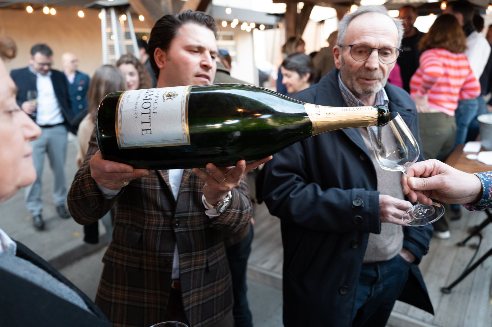 Man in plaid jacket pours champagne from large bottle into glass at outdoor gathering. Background shows people socializing, warm lighting.