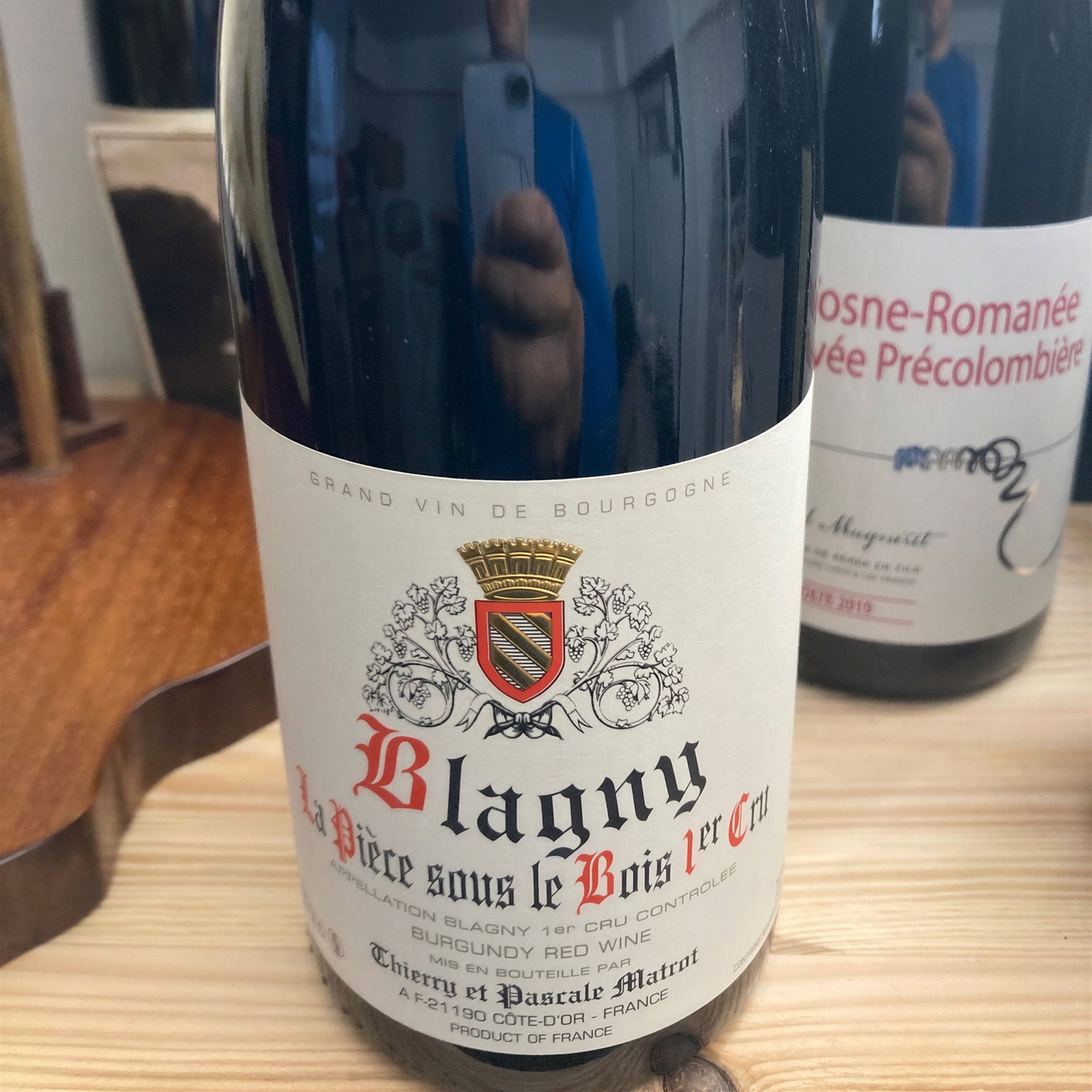 Close-up of a 2019 Blagny wine bottle with a detailed label. Another bottle is partially visible. Reflection of a person taking a photo.