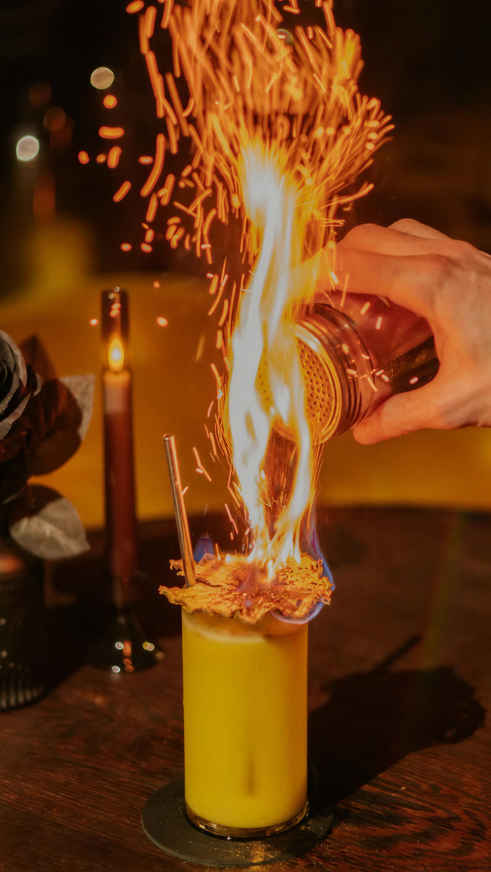 A hand sprinkles spices on a flaming yellow cocktail with a straw. A lit candle and a dark setting create a warm, dramatic atmosphere.