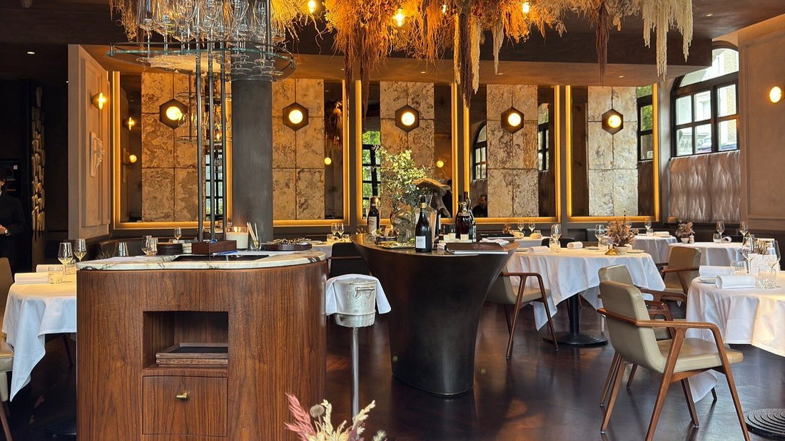 Elegant restaurant interior with white tablecloths, wine bottles, and warm lighting. Wooden accents and dried floral decor create a cozy atmosphere.