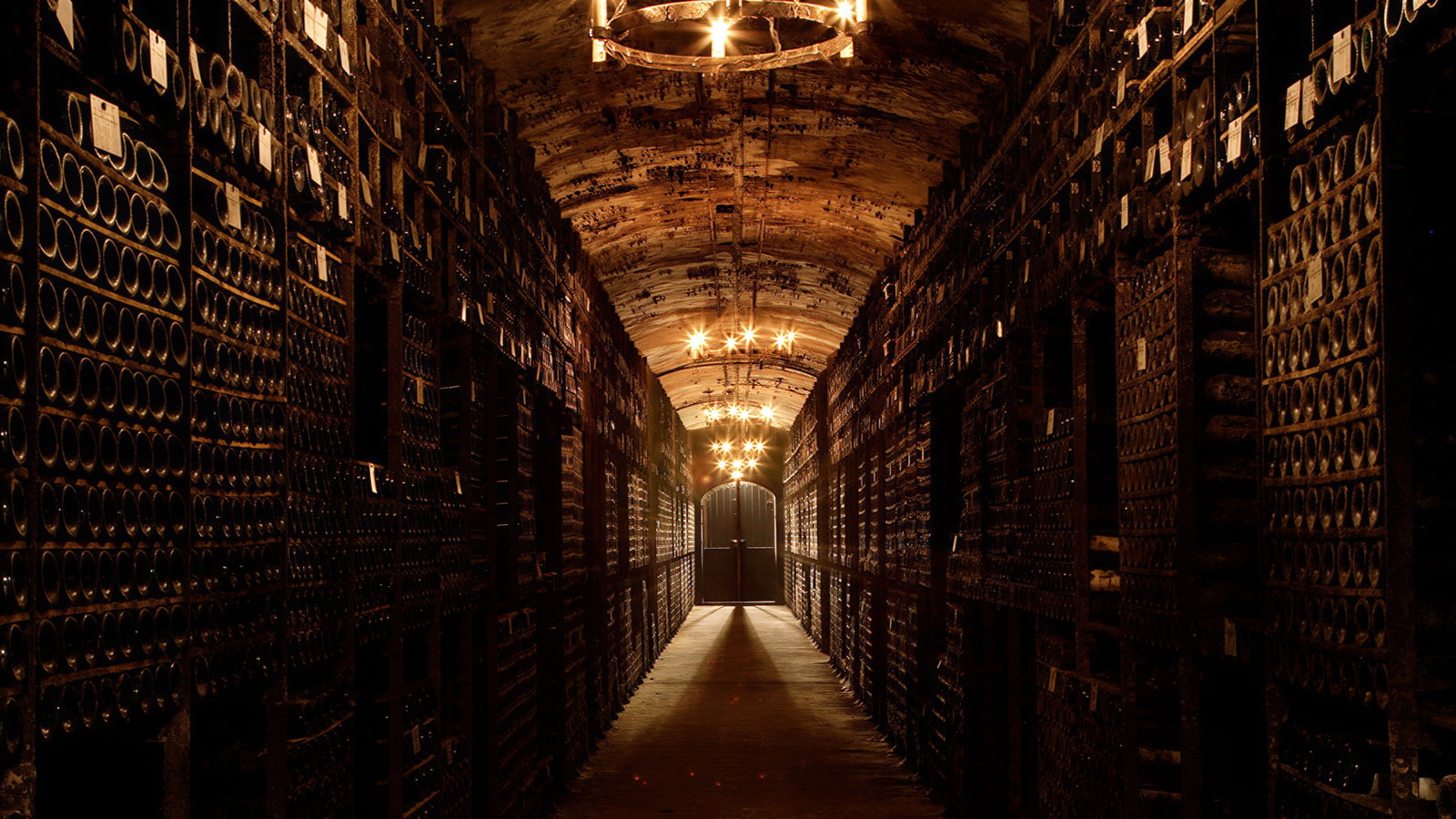 Dimly lit wine cellar with rows of stacked bottles on either side. Warm chandeliers hang from an arched ceiling, creating a cozy ambiance.