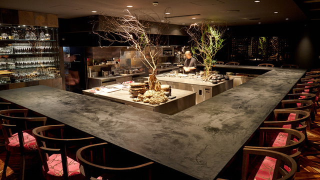 A chef works in a modern kitchen with an L-shaped counter, surrounded by seating. The environment is dimly lit with decorative branches.