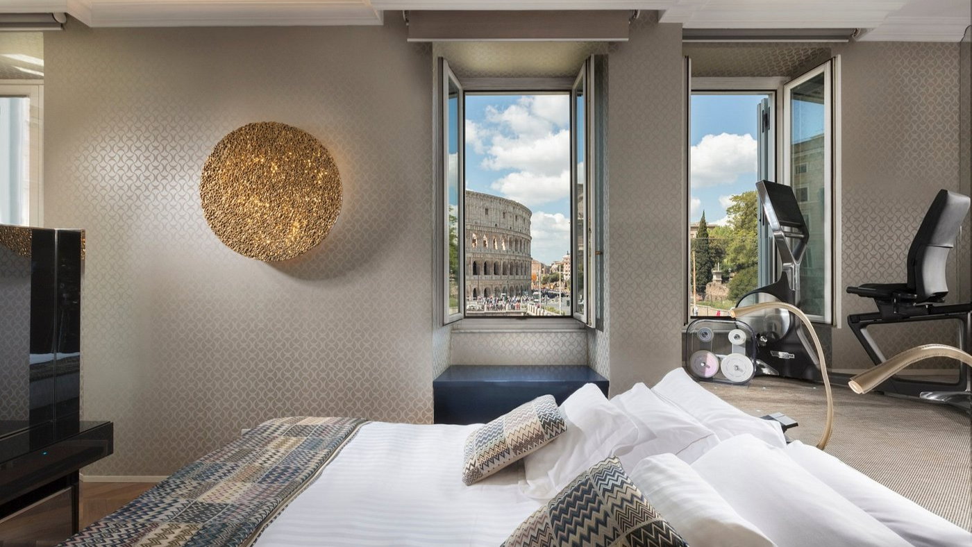 Stylish hotel room with patterned walls, bed, and pillows, exercise bike. Open window view of Colosseum against blue sky, adding elegance.