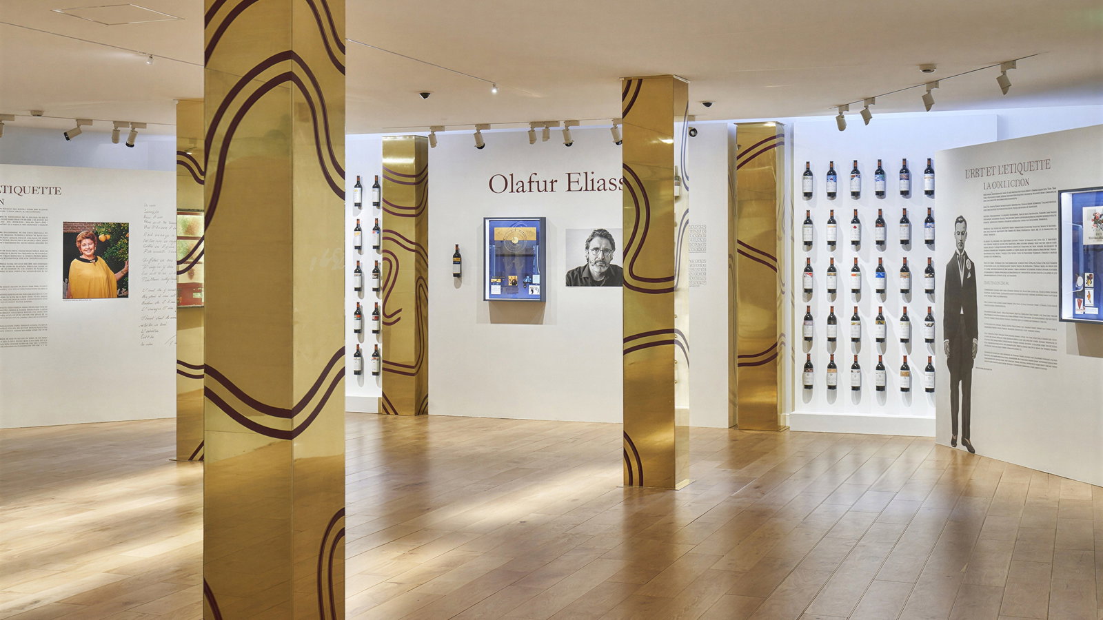Exhibit room with golden columns and wine bottles on display. Text and portraits of a man and woman on white walls. Wood floor. Mood: artistic.