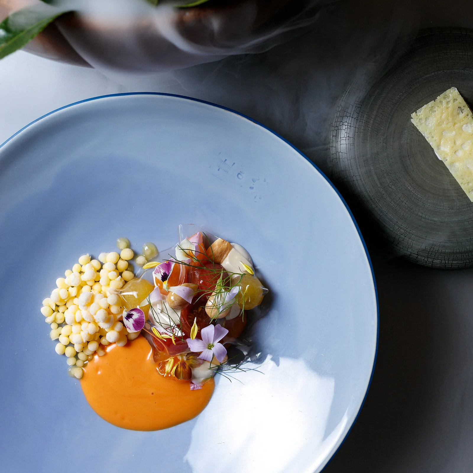 Gourmet dish on a light blue plate with colorful garnish and sauce. Smoke in the background, cheese on a stone plate nearby. Elegant setting.