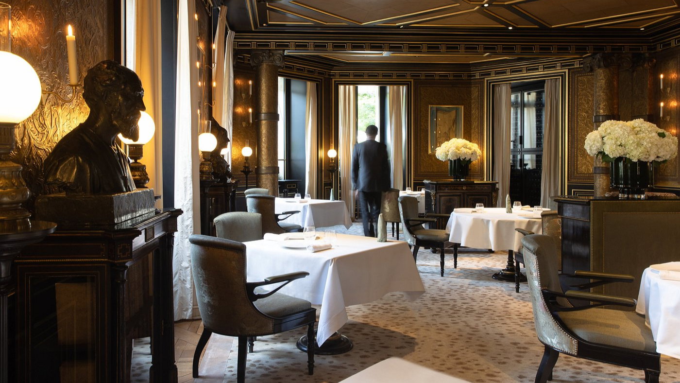 Luxurious restaurant with elegant decor, white tablecloths, floral centerpieces, and a bronze bust. A suited man walks toward large windows.