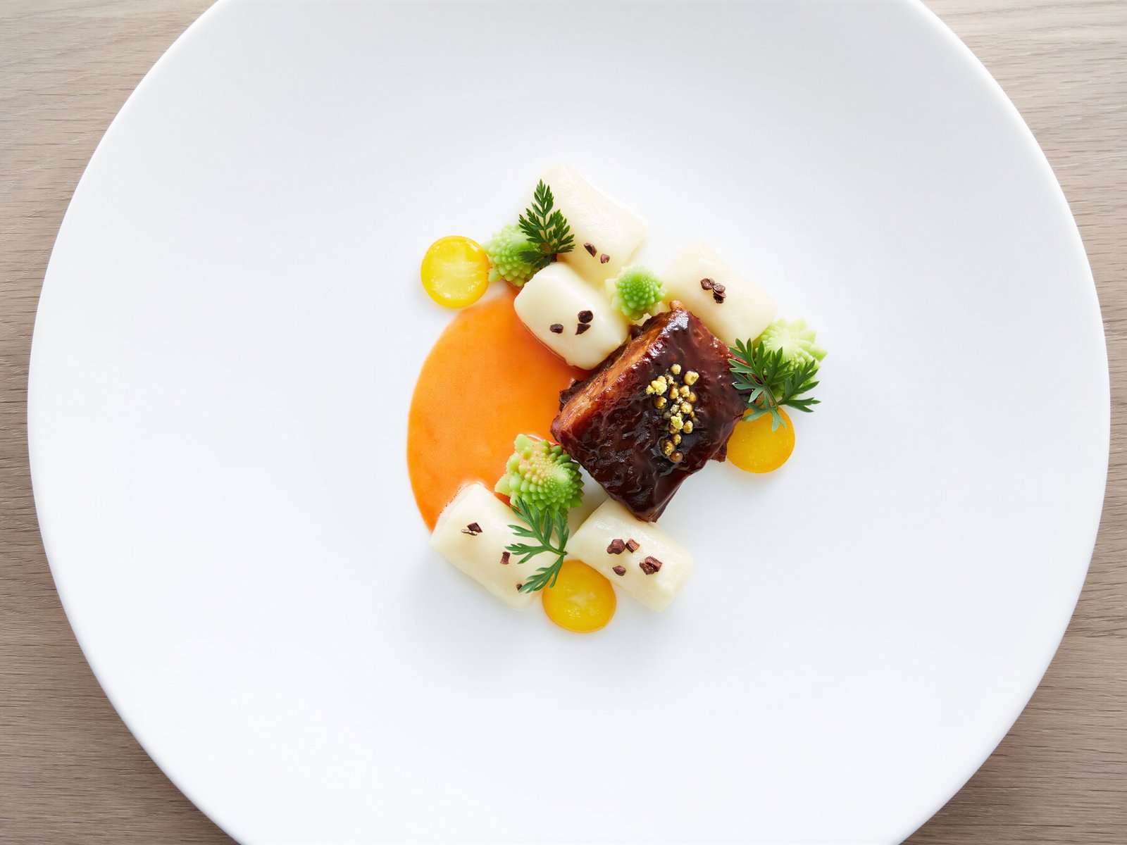 Gourmet dish on a white plate with glazed meat, creamy cubes, orange sauce, and green garnishes. Minimalist presentation on wood table.