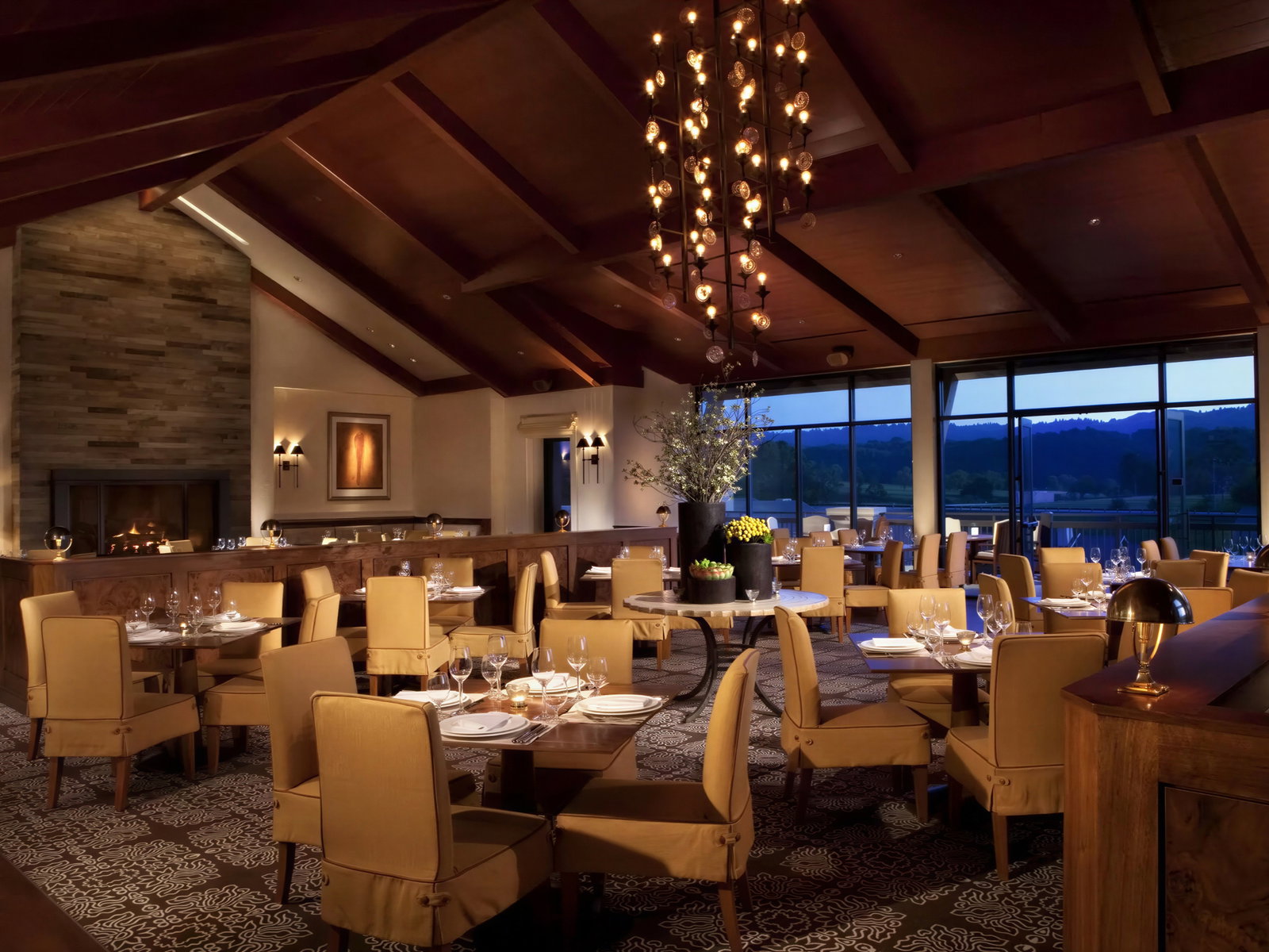 Elegant restaurant with beige chairs, set tables, and a lit fireplace. Large chandelier above, scenic window view, warm and cozy ambiance.
