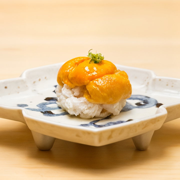 Sea urchin and rice sushi sits on a decorative, white ceramic plate with blue patterns, set on a light wooden surface.