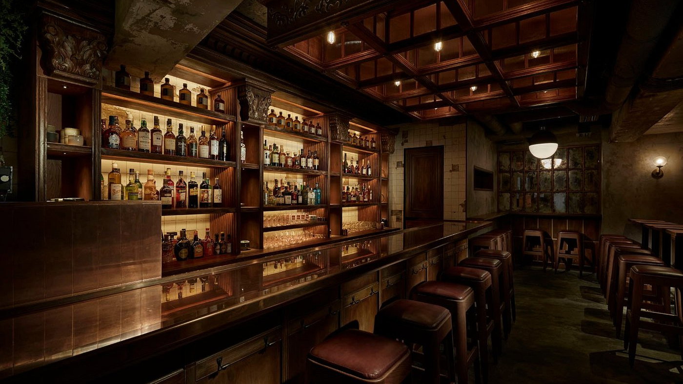 Dimly lit bar with wooden shelves filled with assorted liquor bottles. Brown leather stools line the bar counter in a cozy, warm setting.