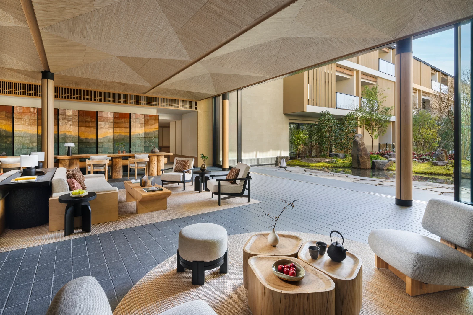 Spacious modern lounge with wooden furniture, gray seats, and a scenic garden view. Warm tones and large abstract wall art enhance calm ambiance.