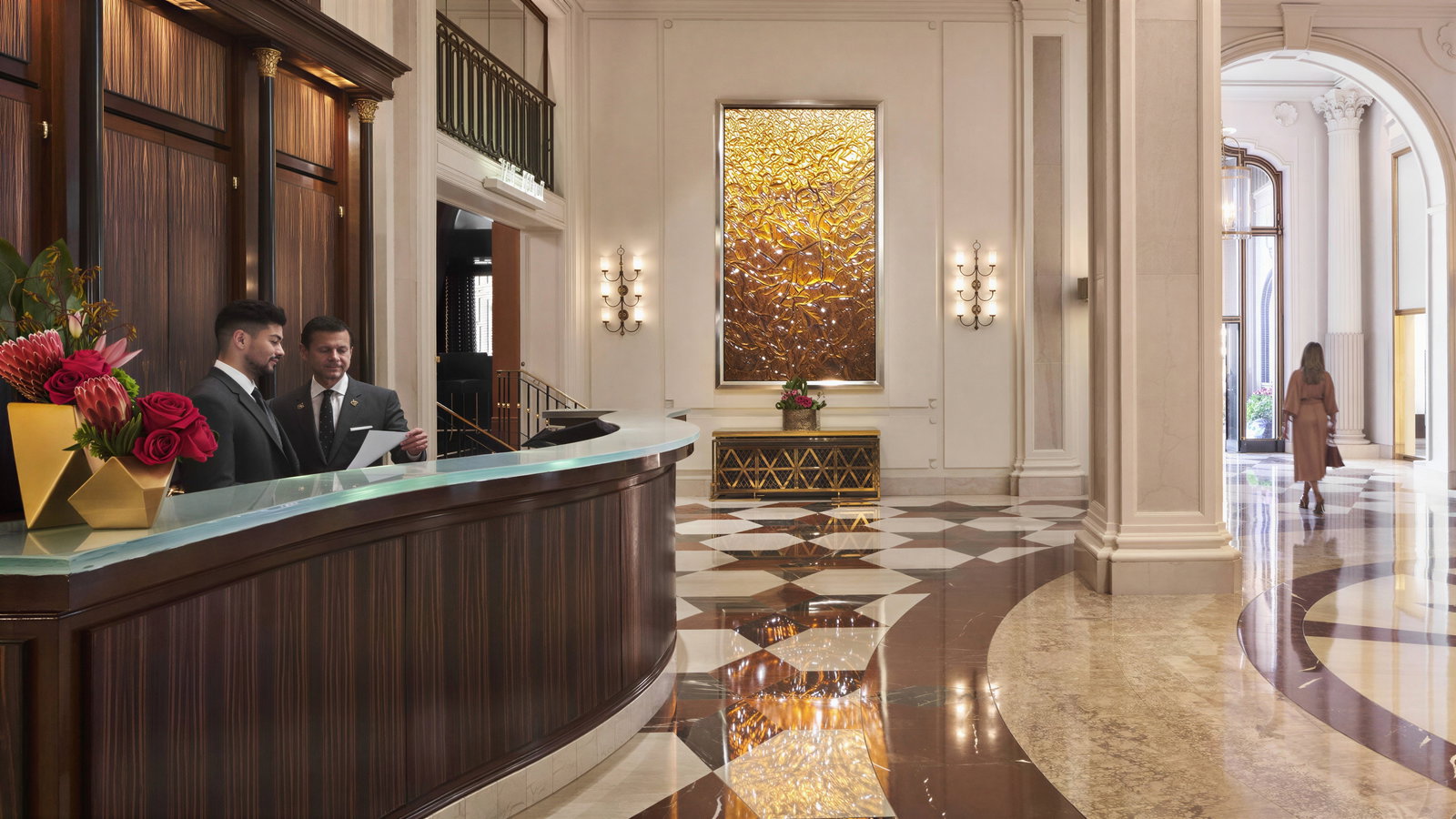 Hotel lobby with two suited men at a reception desk reviewing papers. A woman walks down a marble corridor. Warm lighting, elegant decor.