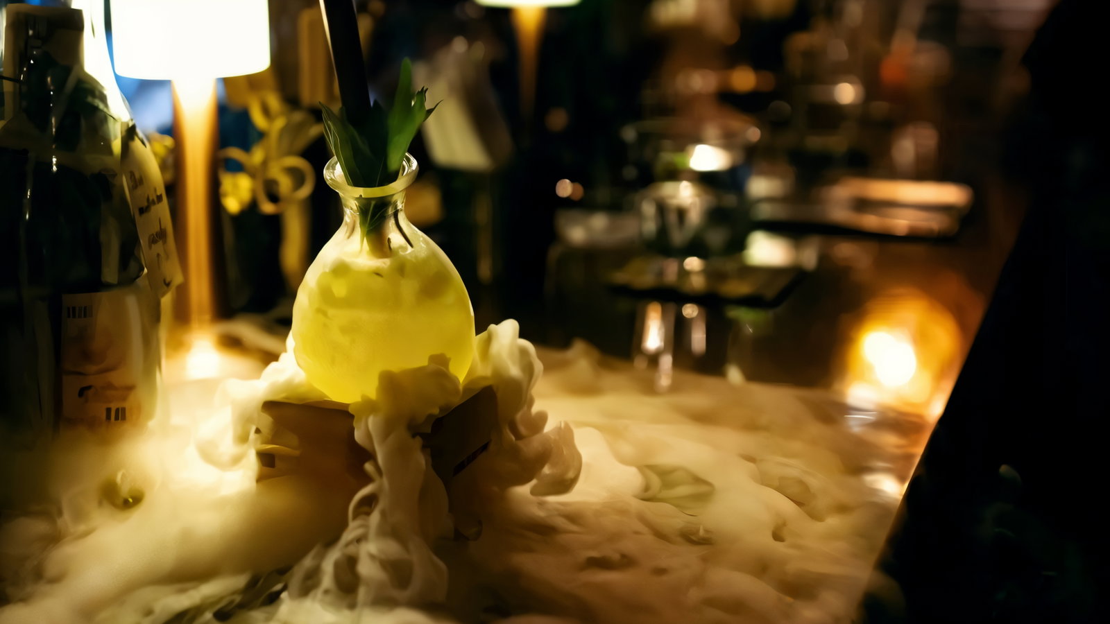 A glowing drink in a glass flask with a straw and leaf garnish sits on a smoky bar, surrounded by warm, dim lights and a cozy atmosphere.