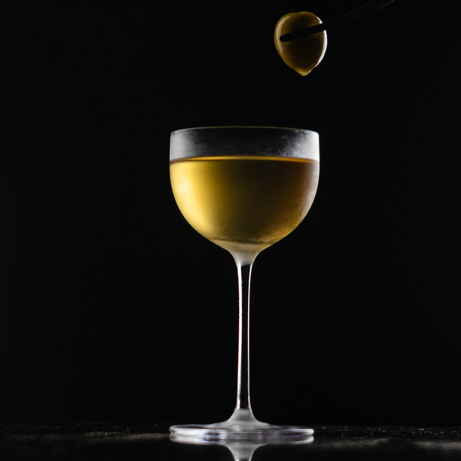 A golden cocktail in a frosted glass on black background. A green olive held by tongs hovers above, suggesting garnish. Elegant, minimalistic.