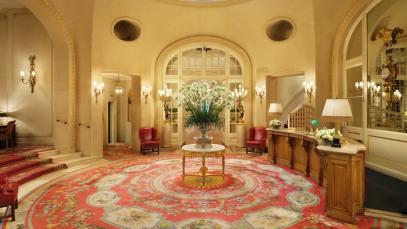 Elegant hotel lobby with ornate decor, red floral carpet, and symmetrical lighting. Central table holds large bouquet. Majestic ambiance.