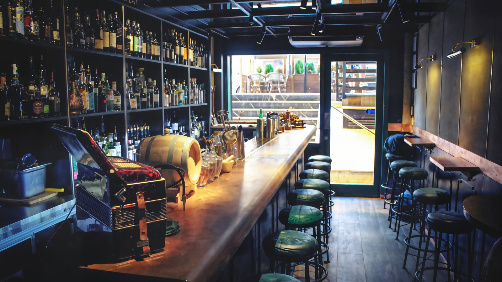Cozy bar with a long countertop lined with green stools. Shelves filled with bottles. Sunlight streams in through the glass door.