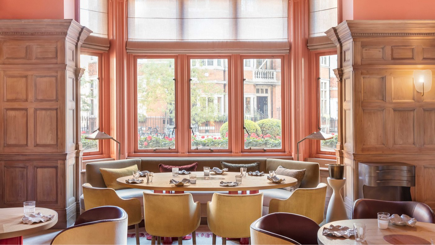 Cozy restaurant interior with wooden panels, colorful chairs, and round tables set for dining. Large windows reveal a leafy street view.