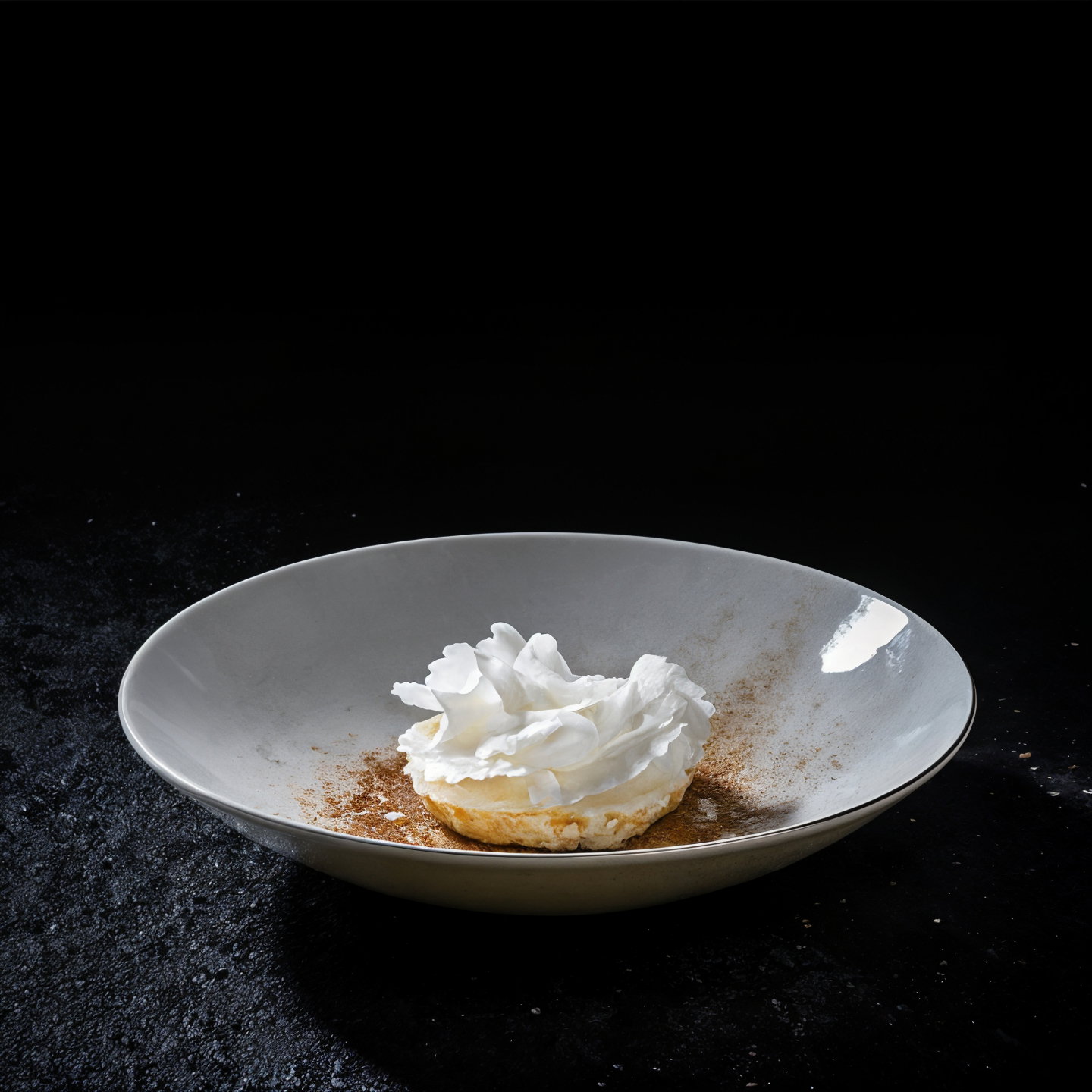 Dessert on a white plate, topped with whipped cream, surrounded by cinnamon powder, set against a dark background.